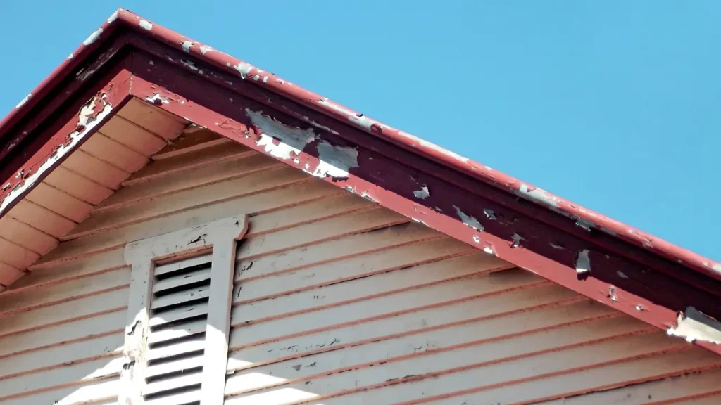 Peeling paint on an older home’s exterior siding and eaves under a clear blue sky